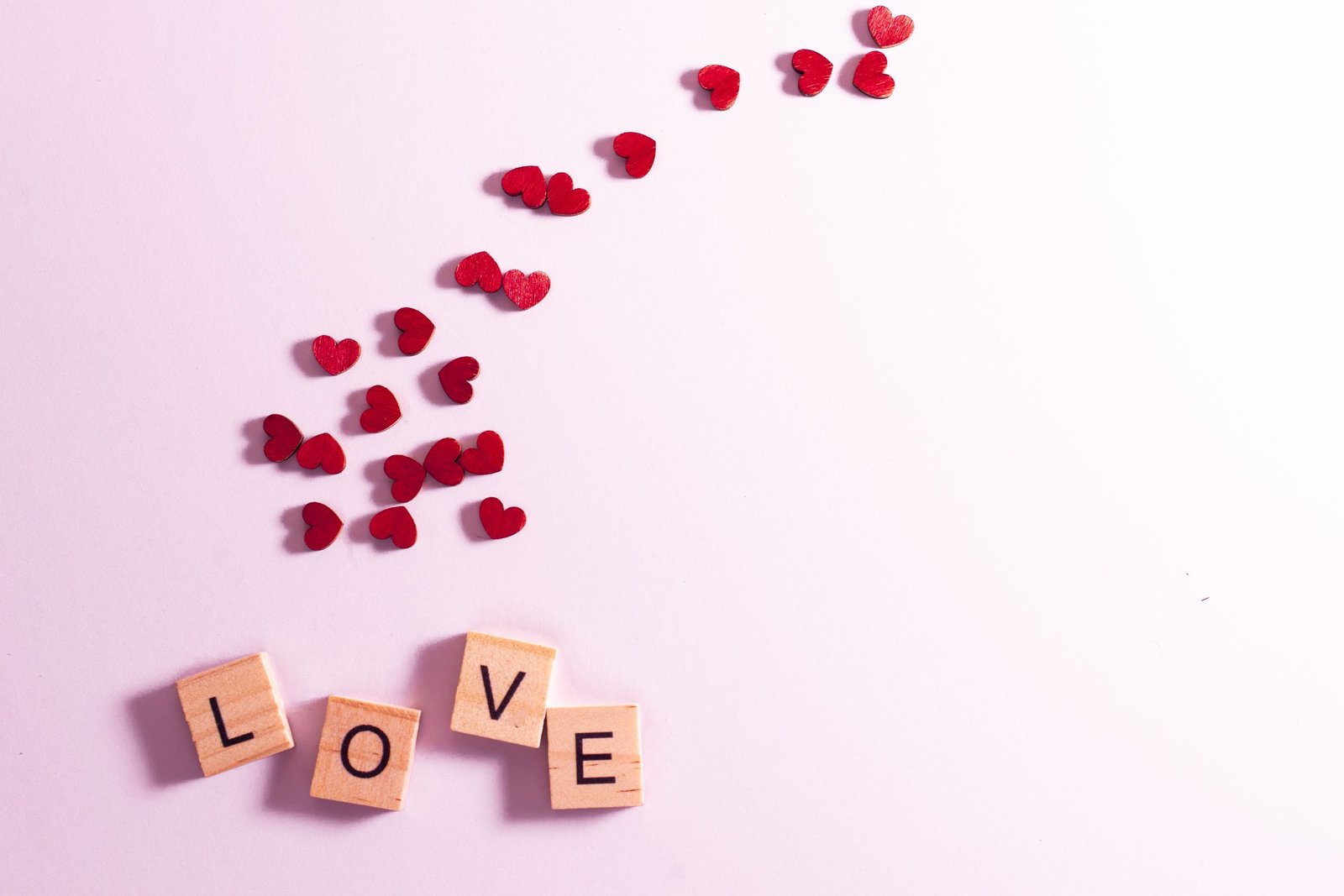 Wooden Scrabble tiles spell 'love' surrounded by red hearts on a pink background, ideal for Valentine's themes.